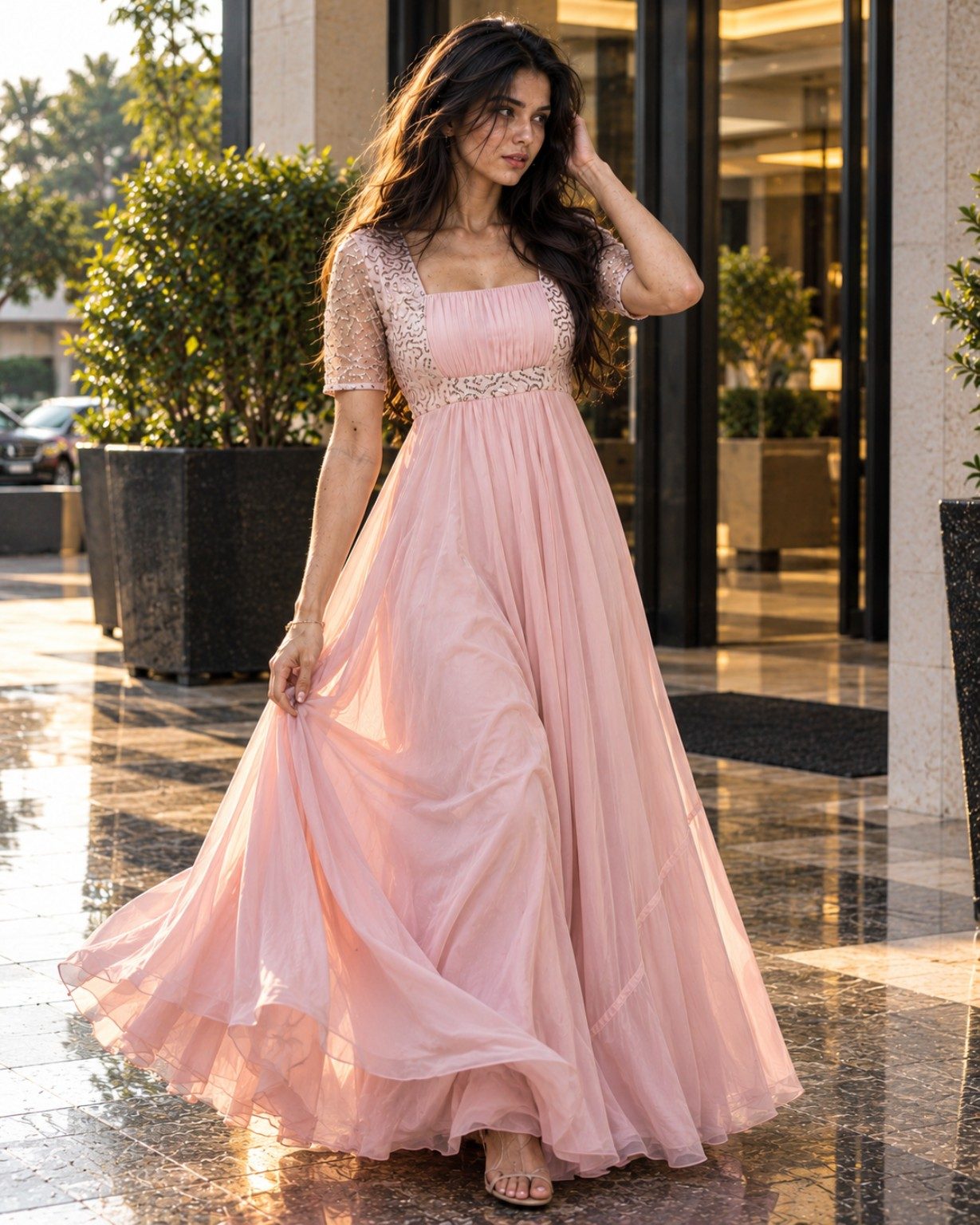 Nyra Voss standing in a flowing blush-pink embellished maxi dress in a sunlit luxury hotel entrance.